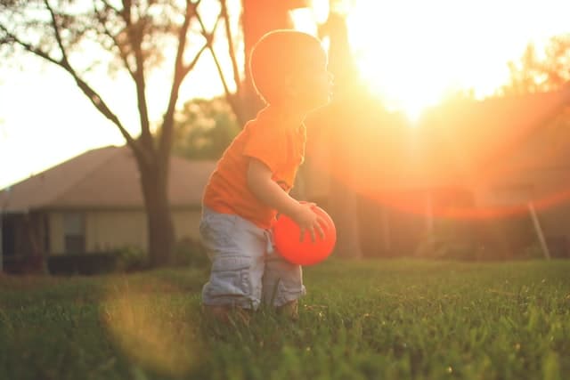 Young boy in yard near house with trees holding orange balloon with sunshine behind him