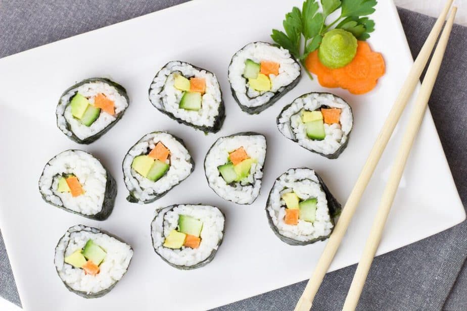 Sushi made with vegetables and rice arranged on white rectangle plate with a pair of bamboo chopsticks and garnished with carrot slice as a flower with wasabi on top