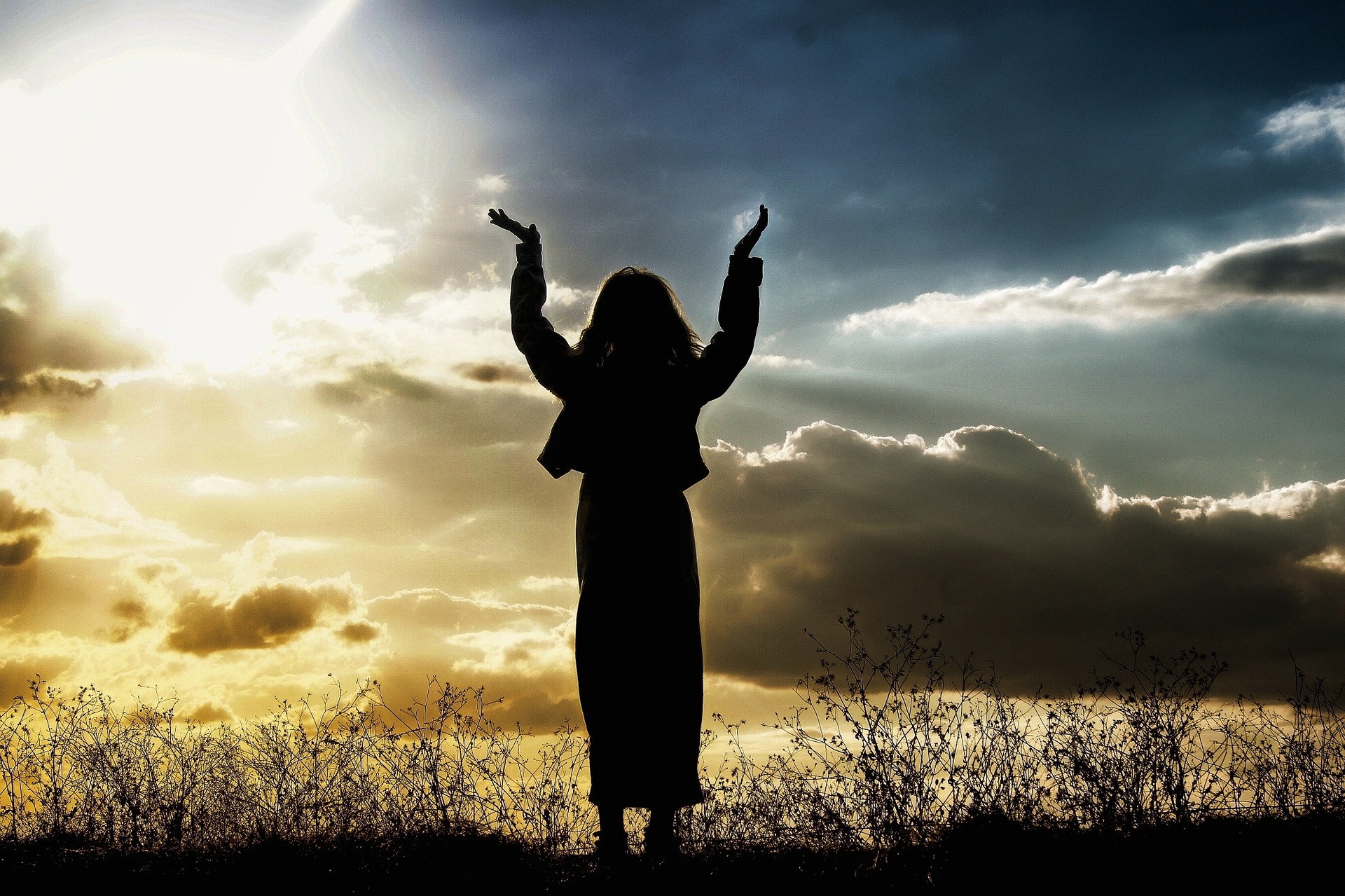 Woman in silhouette standing outdoors with hands raised sky with clouds and sun