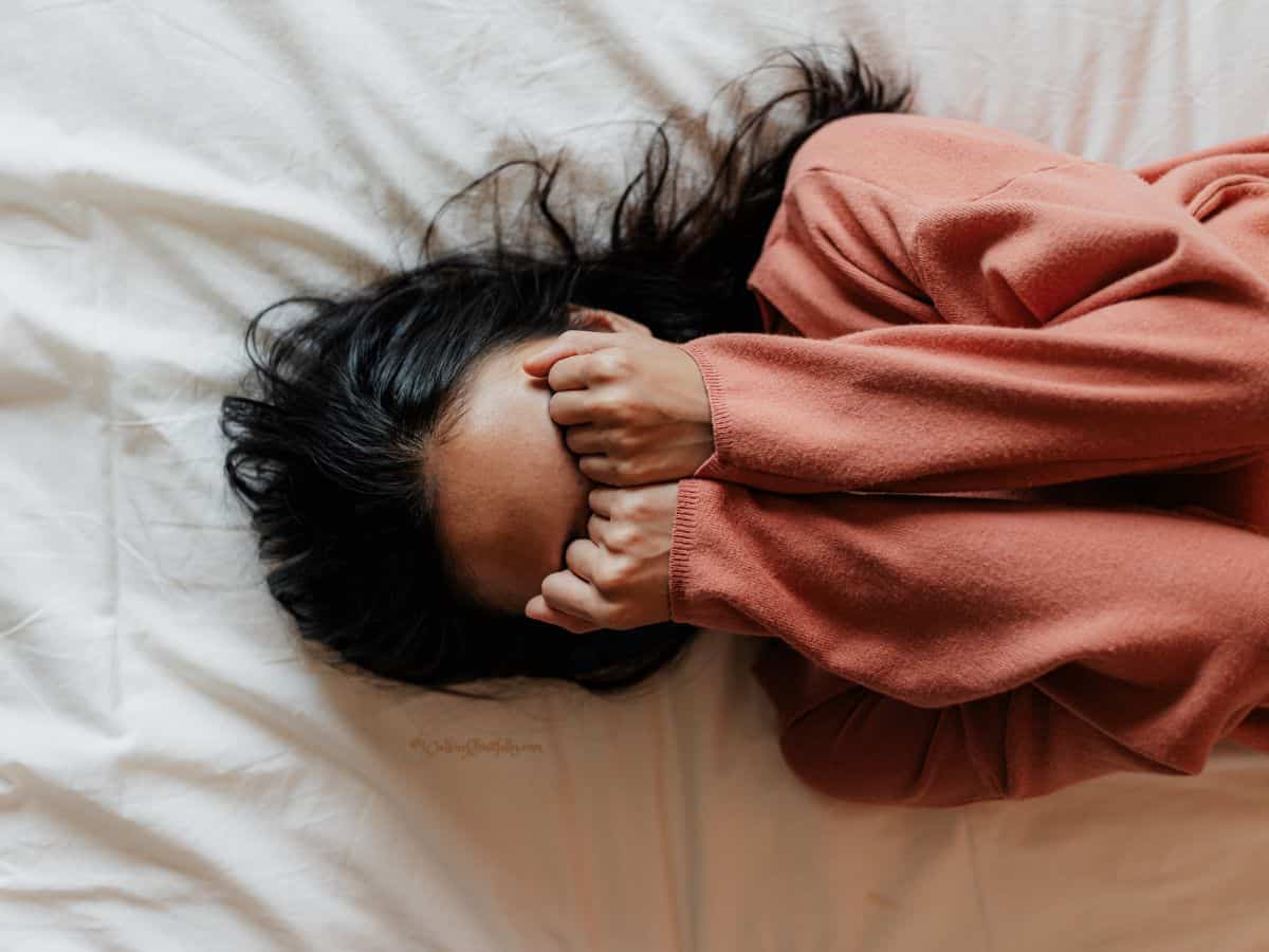 Woman lying on white sheet wearing an orange sweater with her hands covering her eyes