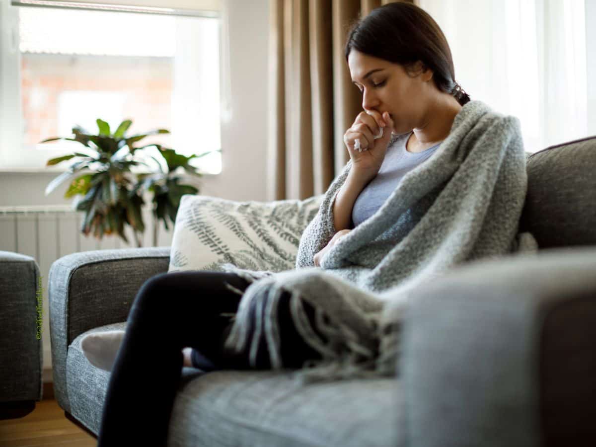 Woman sitting on sofa wrapped in blanket. Tissue in her hand held to her mouth.