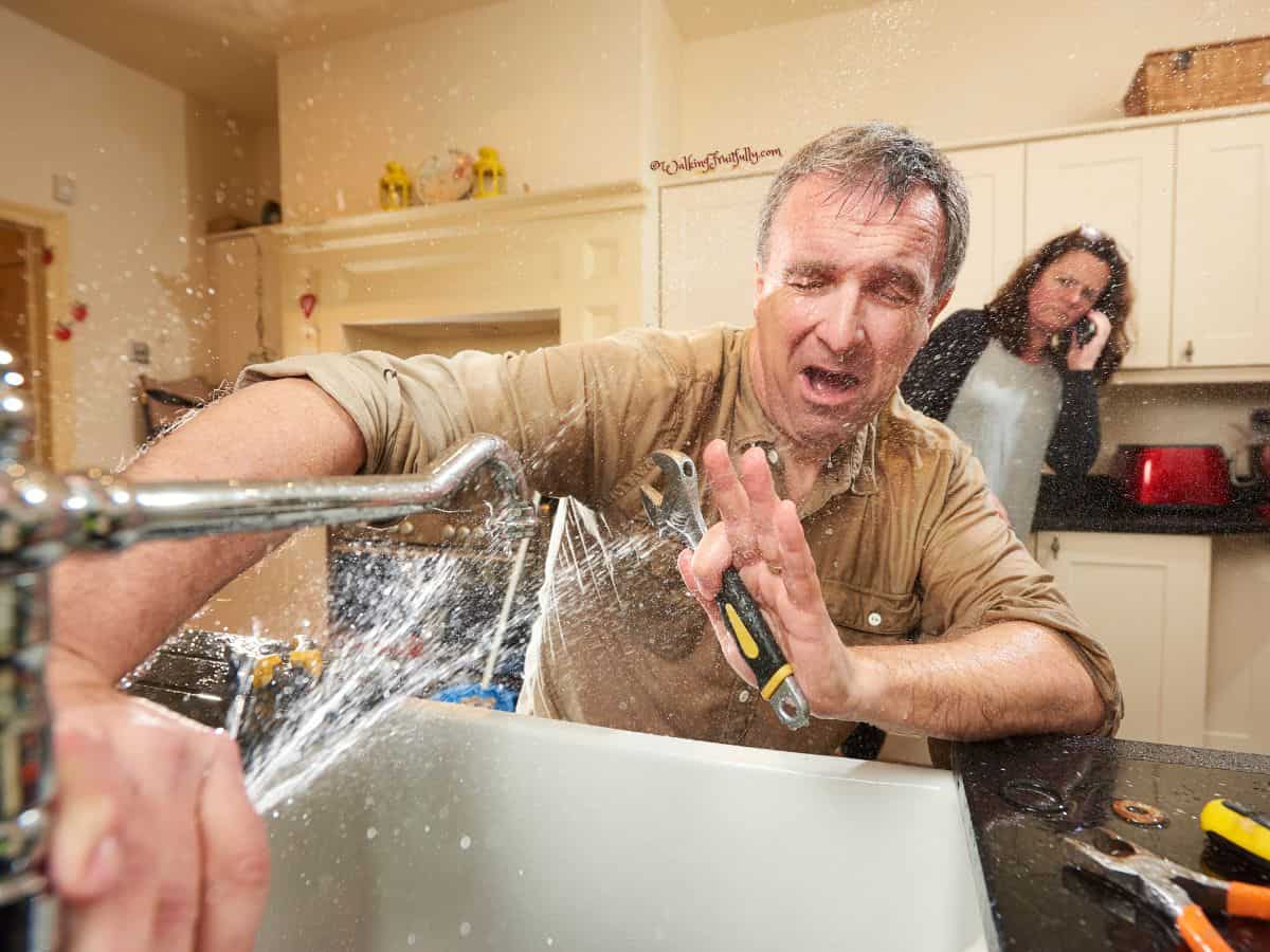 Man with a wrench being sprayed by a leaking faucet while woman in background is on the phone