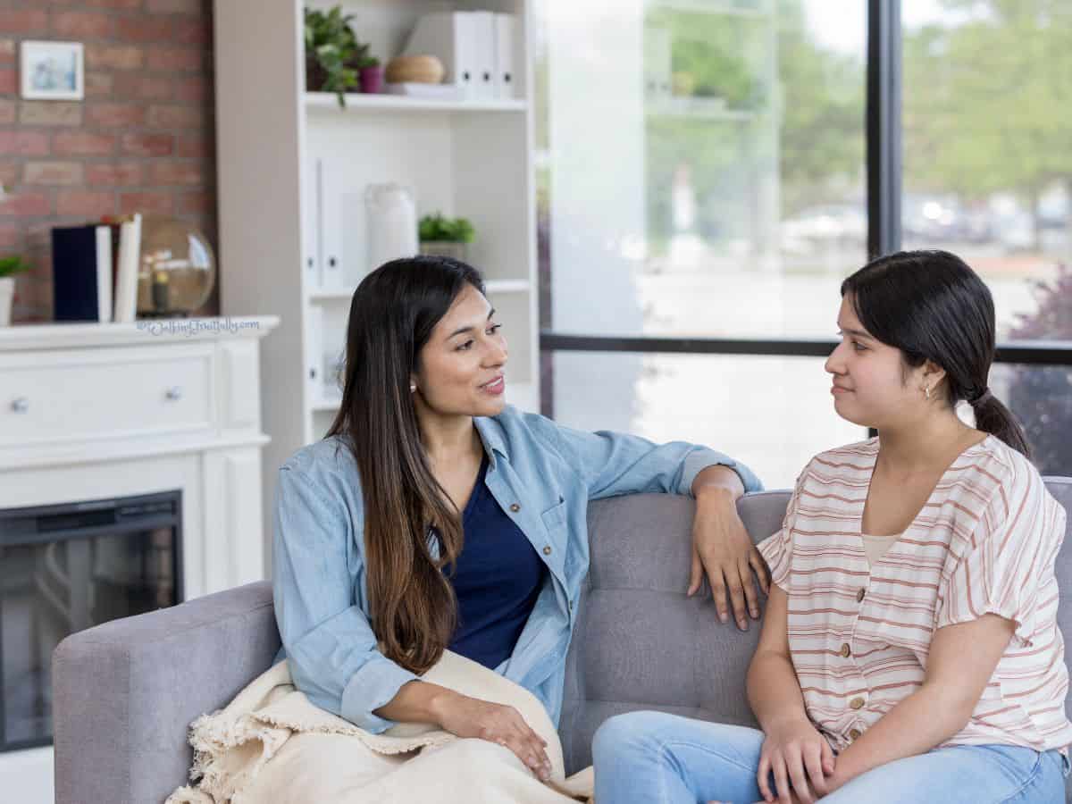 Adult mom and teen girl sit on sofa talking together