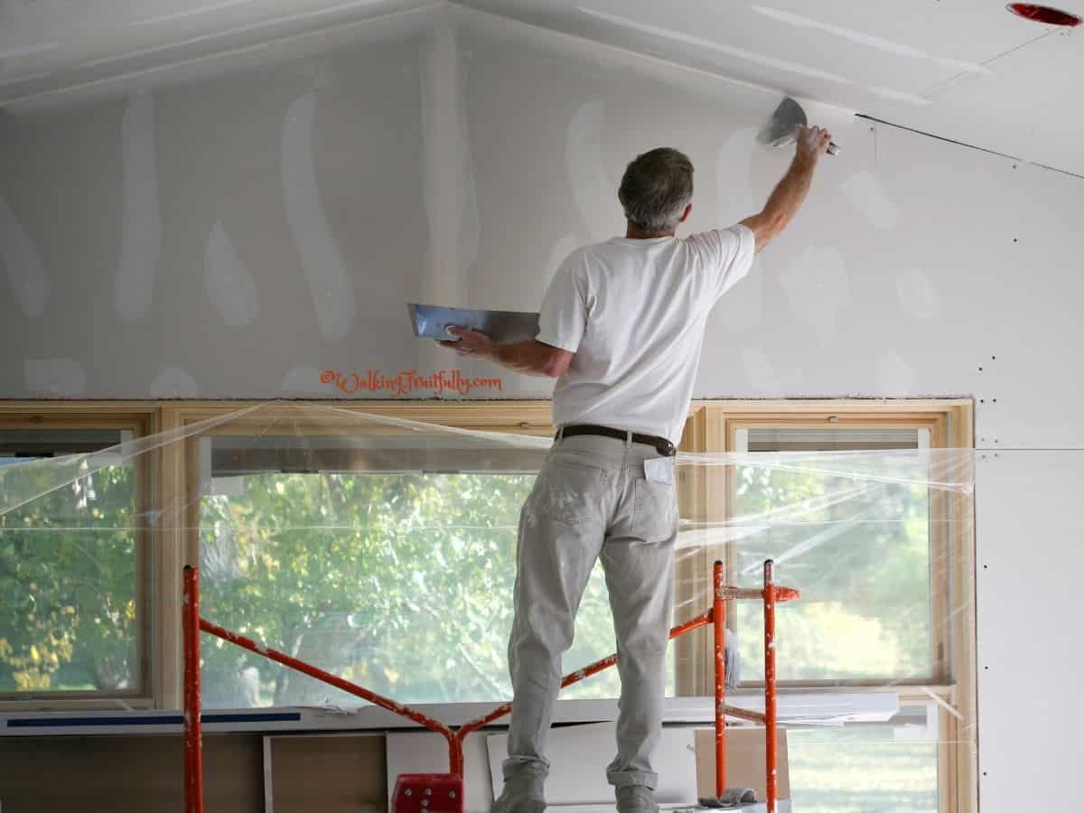 Man on red metal scaffolding applying mud to freshly taped drywall